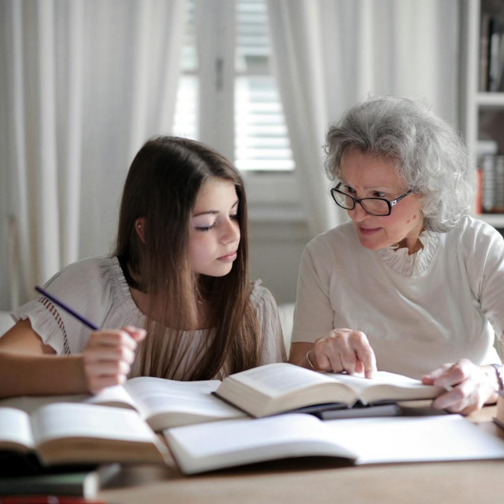 A grandmother helps her teenage granddaughter with homework, surrounded by books indoors.