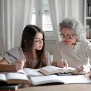 A grandmother helps her teenage granddaughter with homework, surrounded by books indoors.